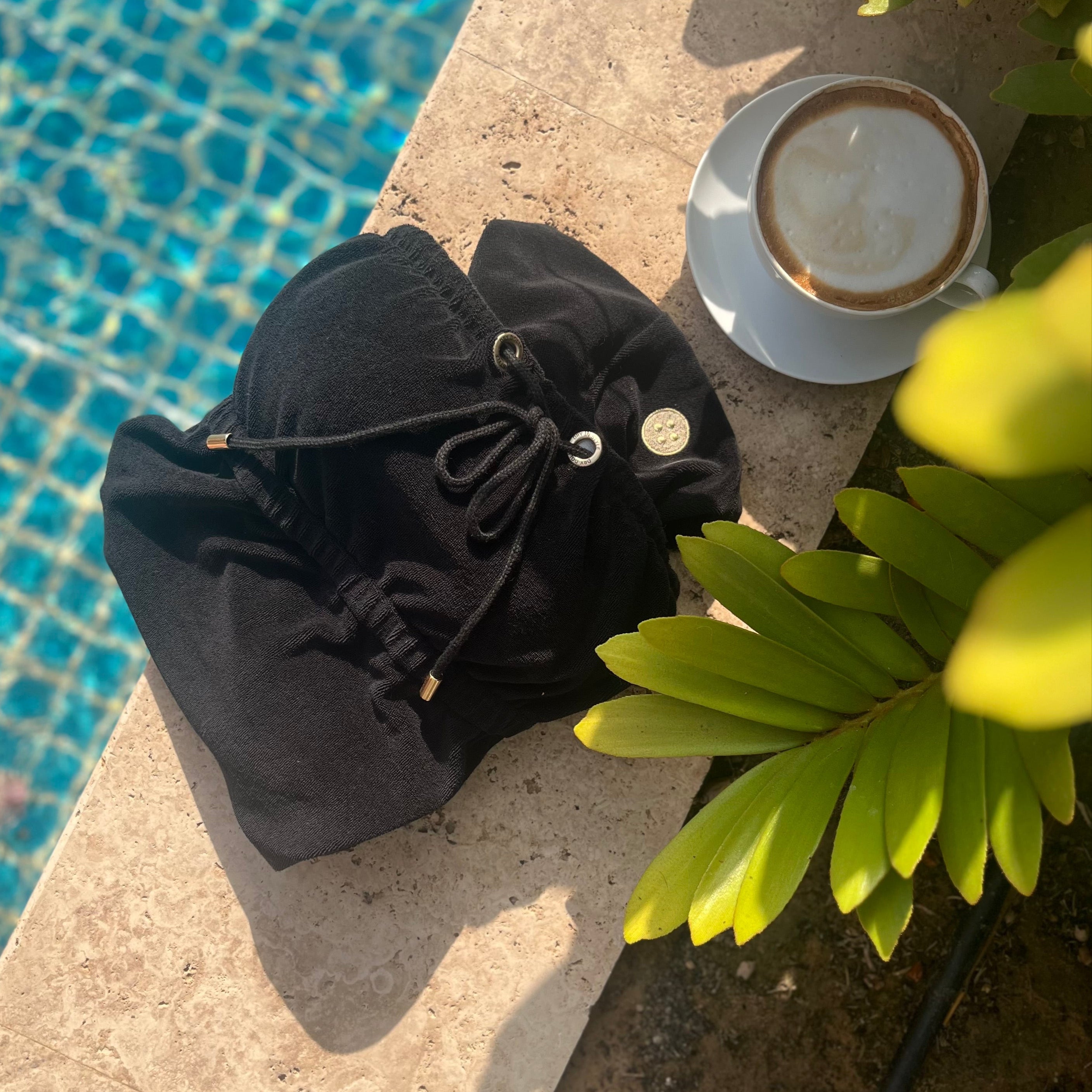 Black Classic Dry Dolly towel dress laid out by a swimming pool beside a coffee cup, capturing relaxed luxury and stylish comfort