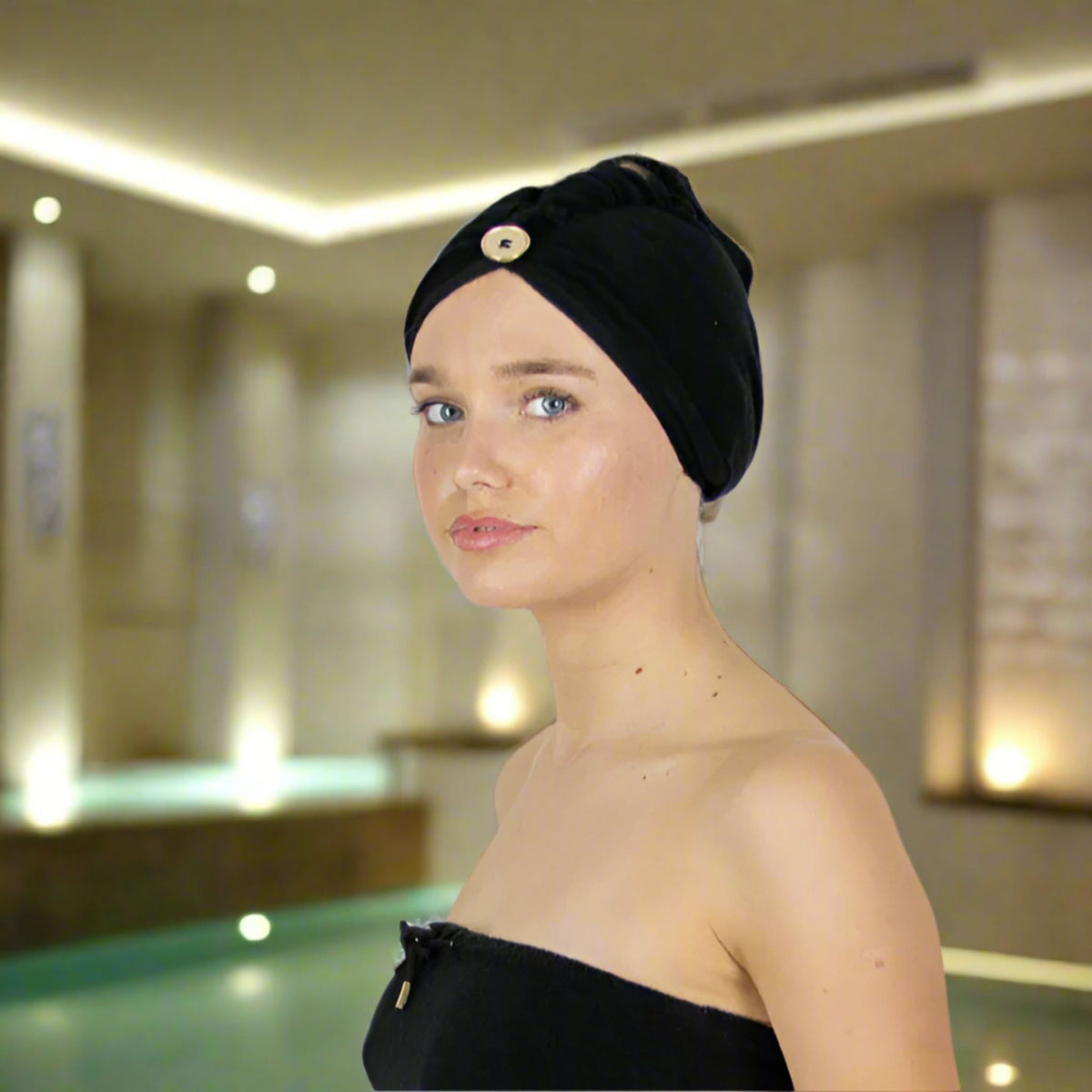 A woman wearing a black towelling turban-style head wrap with a decorative gold button, photographed indoors in a softly lit spa-like environment.